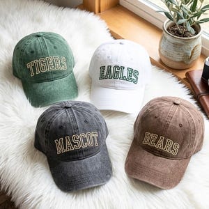 Puede incluir: Cuatro gorras de béisbol en verde, blanco, gris y marrón. Cada gorra tiene un nombre de equipo bordado en un color contrastante: "TIGERS", "EAGLES", "MASCOT" y "BEARS". Las gorras están sobre una superficie blanca y esponjosa.