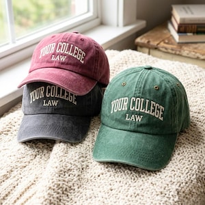 Peut inclure: Trois casquettes de baseball bordeaux, anthracite et verte. Chaque casquette porte l'inscription "YOUR COLLEGE LAW" brodée en blanc. Les casquettes sont posées sur une surface texturée de couleur crème, près d'une fenêtre.
