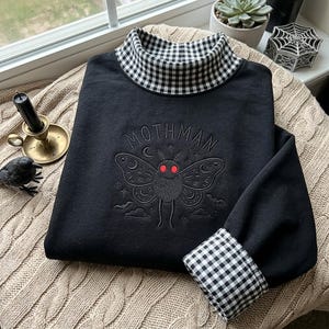 Può includere: Una felpa nera a maniche lunghe con collo alto e polsini a motivo vichy bianco e nero. Sul petto è presente un ricamo raffigurante una falena con occhi rosso vivo e la scritta MOTHMAN arcuata nella parte superiore.