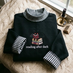 Può includere: Una felpa nera a maniche lunghe con collo alto e polsini in un motivo a quadretti vichy bianco e nero. Il petto presenta un ricamo di una falena su una pila di libri, con il testo reading after dark scritto sotto.