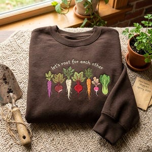 May include: A dark brown crewneck sweatshirt featuring an embroidered design of various colourful root vegetables and the text "let's root for each other" in white script lettering across the centre of the chest area.