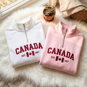 Pode incluir: Duas camisolas, uma branca e outra rosa, com gola com fecho de correr. Cada uma apresenta a palavra "CANADA" em vermelho, a bandeira canadiana e "EST. 1867". As camisolas estão sobre uma superfície branca fofa.