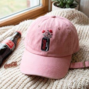 May include: A light pink baseball cap featuring an embroidered graphic of a black Coca-Cola Zero soda can with red flowers emerging from the top. The cap has a curved brim, stitched eyelets, and an adjustable strap with a metal buckle at the back.