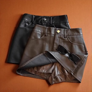 May include: Two faux leather mini skirts, one black and one brown, are displayed. The skirts have a classic five-pocket design with a button closure. A pair of black sunglasses rests on the brown skirt. The skirts are on a brown surface.