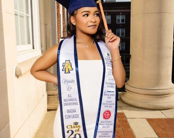 May include: A graduate in a white dress and navy blue cap poses outdoors. She wears a sash with text including "Malaika Lee", "Mechanical Engineering", and "Class of 2025". The sash also lists "Magna Cum Laude" and other achievements.