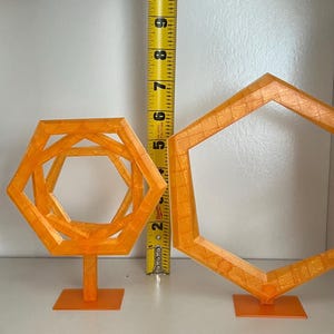 May include: Two orange geometric sculptures, one a nested hexagon design, the other a single hexagon. The nested sculpture is approximately 6 inches tall. Both are on square bases, set against a white background.