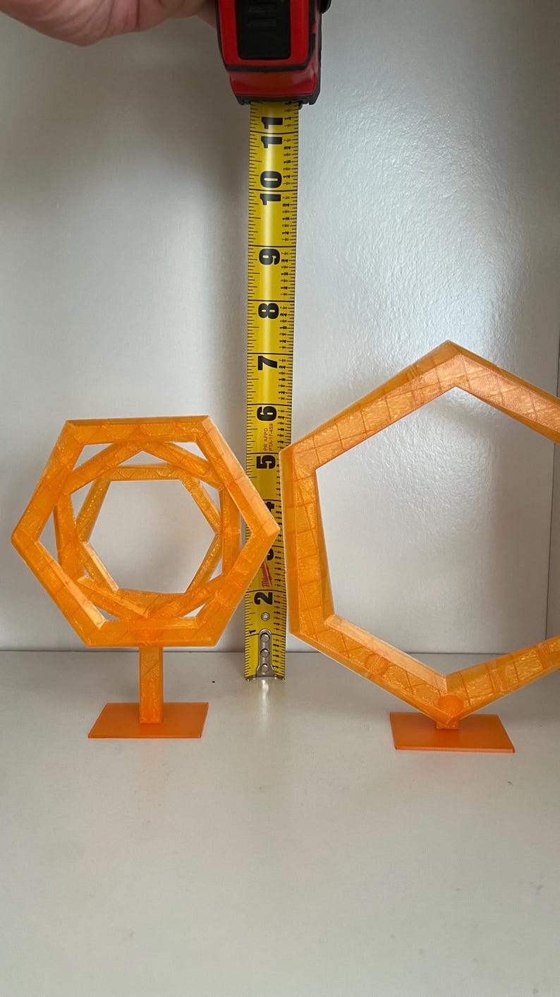 May include: Two orange geometric sculptures, one a nested hexagon design, the other a single hexagon. The nested sculpture is approximately 6 inches tall. Both are on square bases, set against a white background.