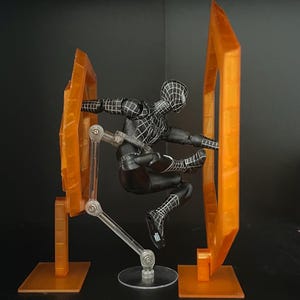 May include: A black Spider-Man action figure in a dynamic pose, set against a dark background. The figure is detailed with white web patterns. It is positioned between two orange, geometric display pieces, connected by clear support arms.