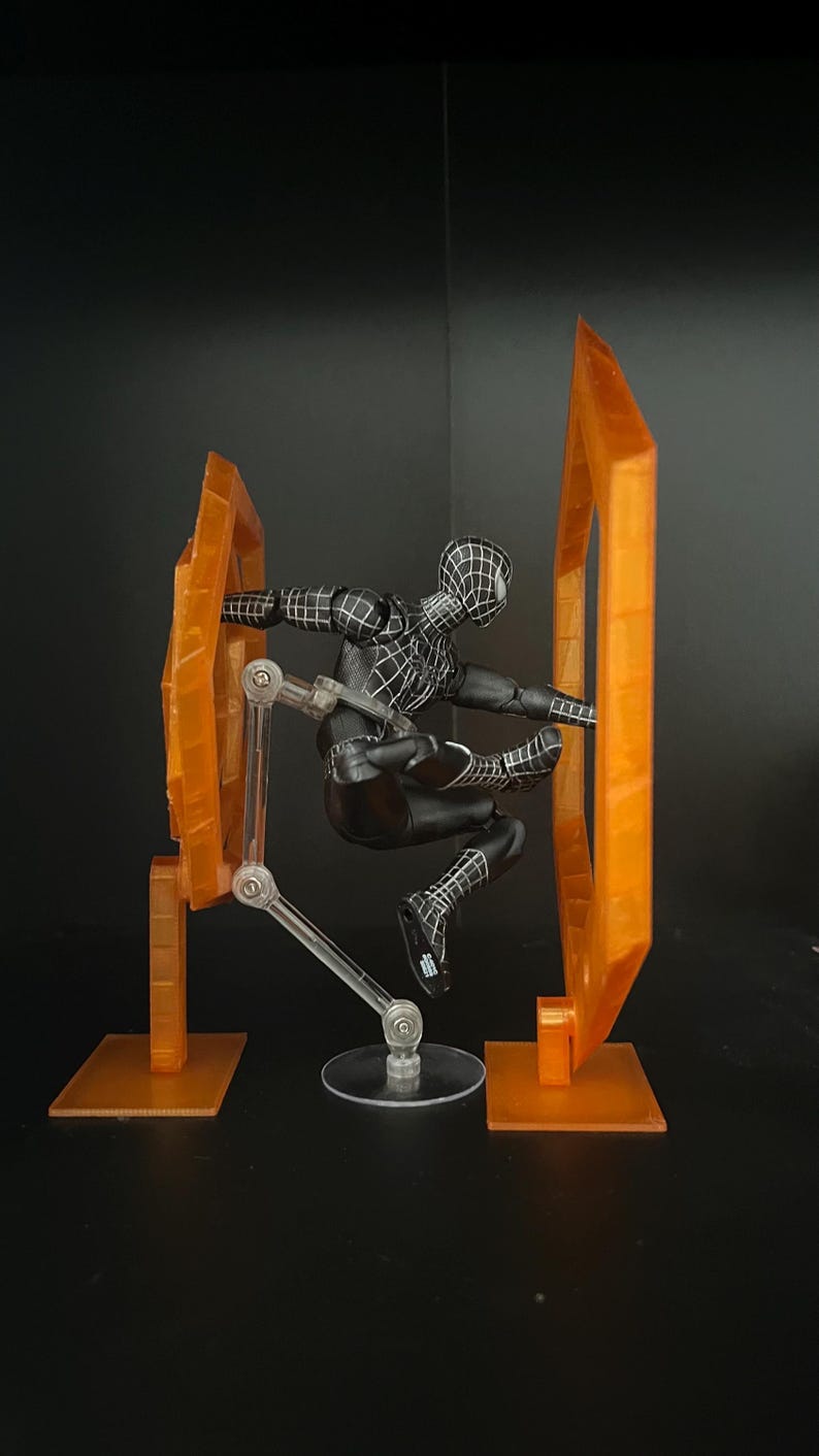 May include: A black Spider-Man action figure in a dynamic pose, set against a dark background. The figure is detailed with white web patterns. It is positioned between two orange, geometric display pieces, connected by clear support arms.