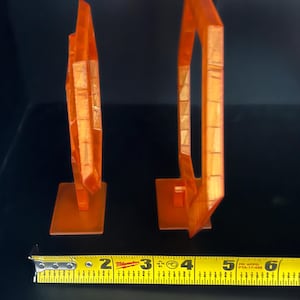 May include: Two orange, geometric sculptures, each mounted on a square base. The sculpture on the left is approximately 3 inches tall, while the one on the right is about 5 inches tall. A measuring tape is in the foreground.