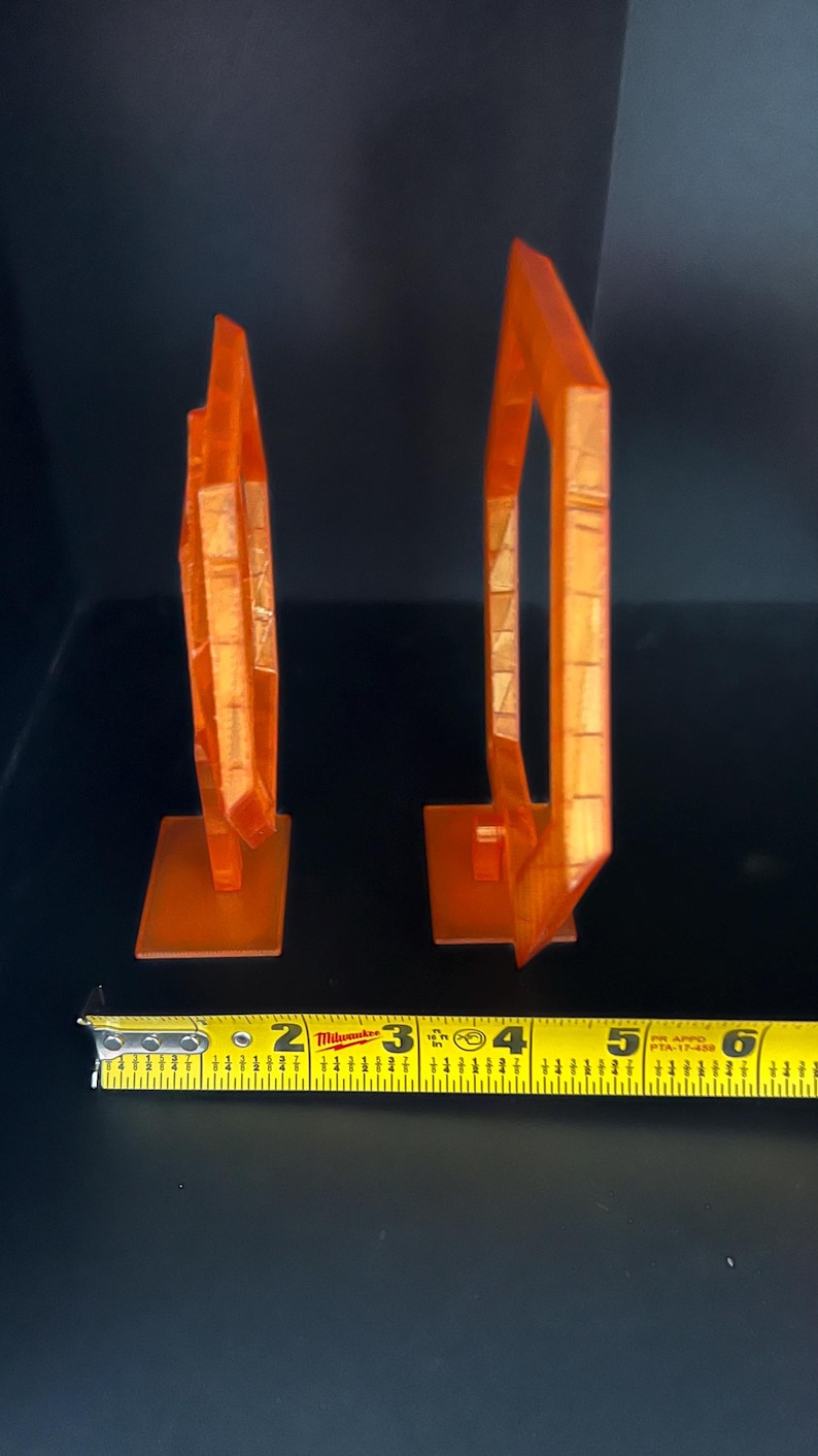 May include: Two orange, geometric sculptures, each mounted on a square base. The sculpture on the left is approximately 3 inches tall, while the one on the right is about 5 inches tall. A measuring tape is in the foreground.