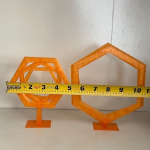 May include: Two orange geometric shapes, one with an interior structure and the other a simple hexagon. The first shape is approximately 4 inches wide, and the second is about 6 inches wide. Both are on small stands.
