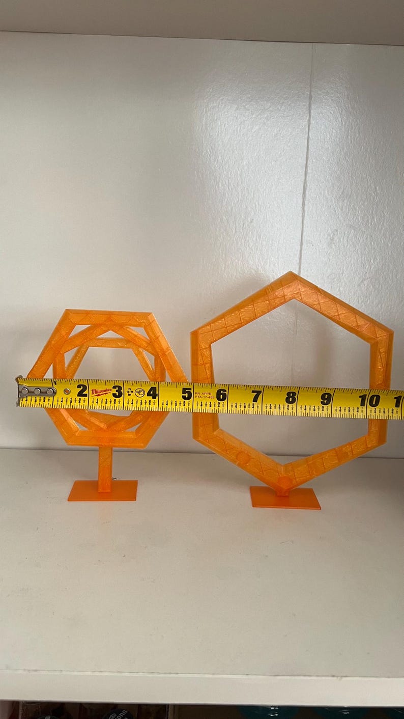 May include: Two orange geometric shapes, one with an interior structure and the other a simple hexagon. The first shape is approximately 4 inches wide, and the second is about 6 inches wide. Both are on small stands.