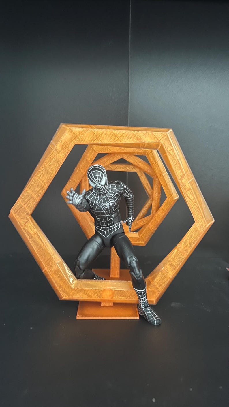 May include: A black and white Spider-Man action figure in a dynamic pose, set against a geometric, orange-colored, hexagonal structure. The figure is detailed with web patterns and stands on a small base.