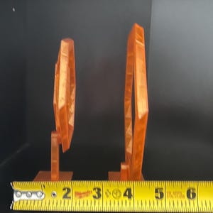May include: Two orange geometric sculptures, one measuring approximately 2 inches tall and the other about 4 inches tall, are displayed against a dark background. The sculptures are made of a translucent material and have a modern design.