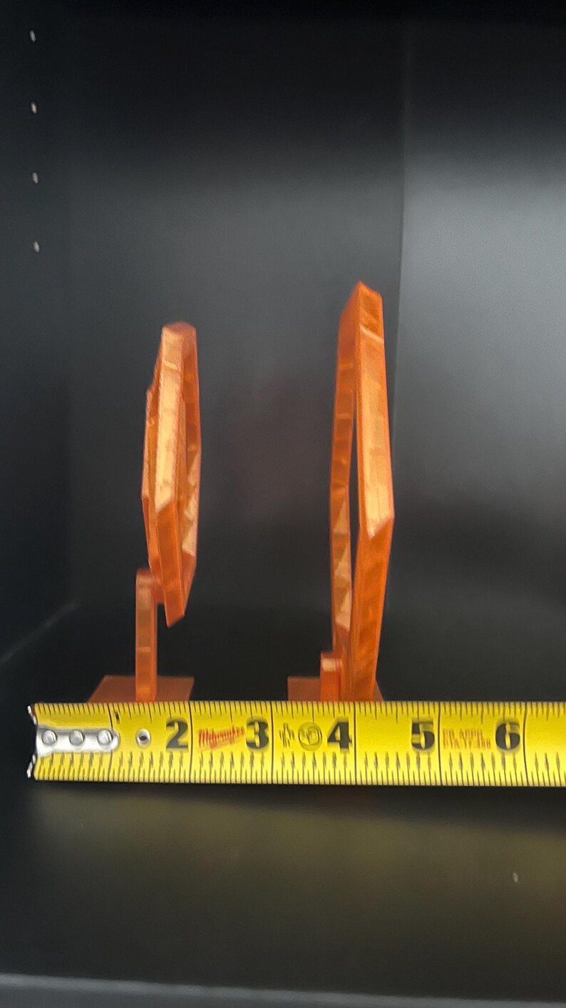 May include: Two orange geometric sculptures, one measuring approximately 2 inches tall and the other about 4 inches tall, are displayed against a dark background. The sculptures are made of a translucent material and have a modern design.