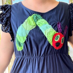 May include: A dark blue dress featuring short ruffled sleeves with eyelet detailing. The front of the bodice displays a large, colorful fabric appliqu&eacute; of a caterpillar with a red head, green body segments, and purple antennae.