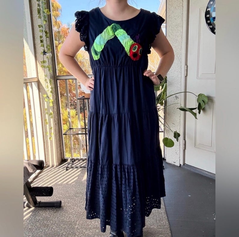 May include: A navy blue tiered maxi dress featuring flutter sleeves with eyelet detailing. The bodice displays a large, colorful fabric appliqu&eacute; of a green caterpillar with a red head. The hem of the skirt also features a matching eyelet fabric panel.