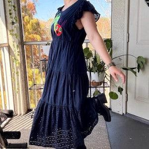 May include: A navy blue midi dress featuring a round neckline, ruffled cap sleeves, and a tiered skirt. The bodice displays a colorful embroidered caterpillar graphic. The bottom tier of the skirt is finished with a decorative eyelet lace trim.
