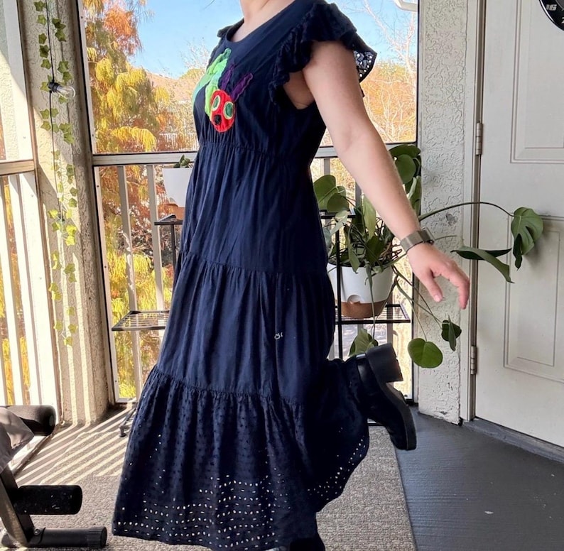May include: A navy blue midi dress featuring a round neckline, ruffled cap sleeves, and a tiered skirt. The bodice displays a colorful embroidered caterpillar graphic. The bottom tier of the skirt is finished with a decorative eyelet lace trim.