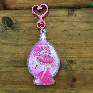 Op de afbeelding: Een roze acryl sleutelhanger met een cartoon illustratie van een meisje in een roze jurk. De sleutelhanger heeft een hartvormige sluiting en staat op een houten achtergrond met roze bloemen en groene bladeren.
