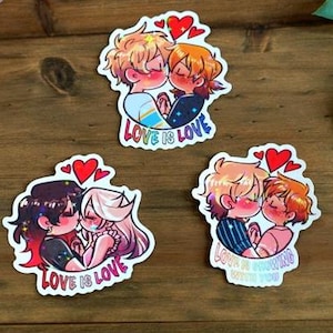 May include: Three stickers depicting cartoon couples kissing, adorned with red heart motifs. The stickers bear the text "LOVE IS LOVE" and "LOVE IS GROWING WITH YOU". The couples have diverse hair colours and wear varied attire. The stickers are presented on a wooden surface.