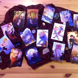 May include: A collection of art prints featuring colorful illustrations of couples in various romantic poses. The prints are arranged on a dark red fabric, with decorative stones and a silver chain scattered around them. The artwork has a fantasy theme.