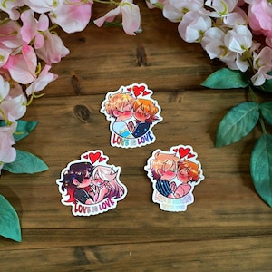May include: Three stickers featuring cartoon couples embracing, each with a heart above them. The stickers have the text "Love is Love". They are displayed on a wooden surface, framed by pink and white flowers.