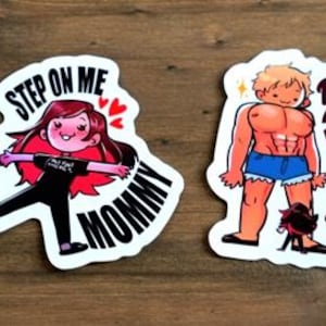 以下が含まれることがあります： 木製の表面に2つの漫画ステッカー。1つは赤い髪の女性と「Step on me Mommy」の文字が特徴です。もう1つは、「Daddy?!」の文字が入った筋肉質な男性を示しています。