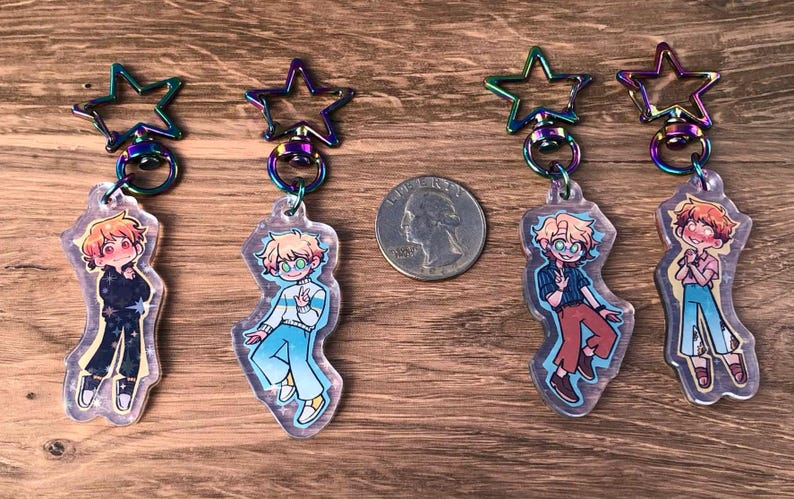 Puede incluir: Cuatro llaveros con cierres en forma de estrella e ilustraciones de dibujos animados de una figura masculina. Los llaveros tienen una forma clara e irregular. Las figuras visten varios atuendos, con diferentes colores de pelo y expresiones. Se incluye una moneda de veinticinco centavos estadounidense para comparar el tama&ntilde;o.