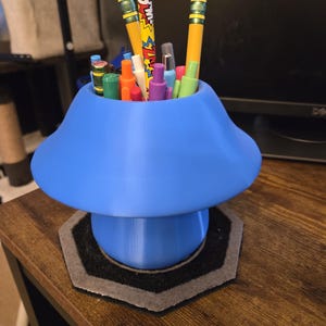 May include: A blue mushroom-shaped desk organizer holding pencils, pens, and markers. The organizer sits on a dark wooden surface with a gray and black hexagonal coaster. The pencils are yellow with pink erasers.