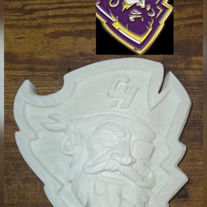 May include: A white, carved wooden pirate emblem with a detailed face, eye patch, and hat. The emblem is shield-shaped. A smaller, colored version of the emblem is in the upper right corner, featuring purple, gold, and white.