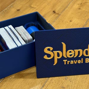 May include: A navy blue travel box for the board game Splendor, with gold lettering. The box is open, revealing stacks of cards, colored tokens, and blue cylinders. The box is on a wooden surface.