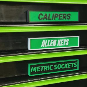 May include: Three green and black labels with white text are displayed. The top label reads "CALIPERS", the middle label reads "ALLEN KEYS", and the bottom label reads "METRIC SOCKETS". The labels are organised on a black surface with green accents.