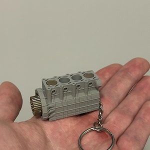 Peut inclure: Un porte-clés miniature gris en forme de bloc moteur avec un porte-clés argenté. Le bloc moteur comporte quatre cylindres et un engrenage à une extrémité. Le porte-clés est tenu dans une main.