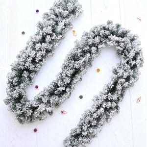 May include: A flocked artificial Christmas garland with a snowy appearance. The garland is green with white flocking, and is arranged in a wavy pattern on a white surface. Small colourful confetti pieces are scattered around the garland.