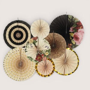 May include: A collection of decorative paper fans in various designs. Includes fans with black and white stripes, gold polka dots, floral patterns, and solid colors. Some fans have gold trim and are arranged on a white background.