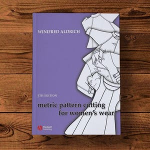 Op de afbeelding: Een boek getiteld "metric pattern cutting for women's wear" van Winifred Aldrich, 5e editie, op een paarse achtergrond. De omslag toont lijntekeningen van dameskleding en het Blackwell Publishing-logo. Het boek ligt op een houten oppervlak.