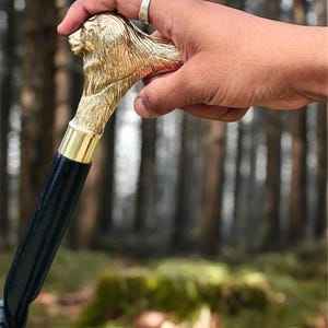 Könnte beinhalten: Ein Gehstock mit einem Griff aus poliertem, goldfarbenem Metall in Form eines Löwenkopfes mit detaillierter Mähnenstruktur. Der Griff ist über eine zylindrische, goldfarbene Metallmanschette mit einem glatten, schwarzen Schaft verbunden.