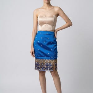 May include: A strapless beige satin corset top paired with a knee-length blue skirt featuring a small diamond pattern and a wide, intricate gold and blue woven border at the hem, including a side slit and matching beige strappy high-heeled sandals.