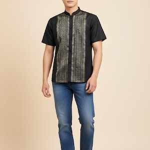May include: A black short-sleeved shirt with a mandarin collar and a gold and black patterned design on the front. The shirt is paired with blue jeans and black trainers. The person is standing in front of a beige background.