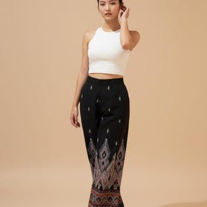 Op de afbeelding: Een vrouw draagt een witte haltertop en een zwarte broek met een decoratief geometrisch patroon in roze, rood en grijs. De broek heeft een wijd been ontwerp. De vrouw draagt beige hoge hakken.