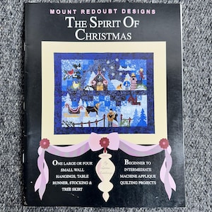 May include: A book cover titled "The Spirit of Christmas" by Mount Redoubt Designs. The cover features a winter scene with a house, trees, and figures. The book offers beginner to intermediate machine applique quilting projects.