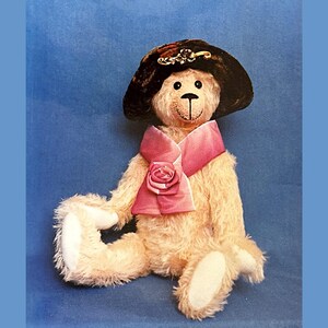 May include: A teddy bear wearing a brown hat with a gold decoration and a pink scarf with a rose. The bear has light tan fur and white paws, set against a blue backdrop. A charming, handcrafted collectible.