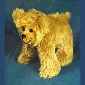 May include: A soft, golden-brown teddy bear with a stitched black nose and smiling mouth. The bear has small, round black eyes and a fluffy, textured fur. It is posed on all fours, with its paws visible, and a small tail.