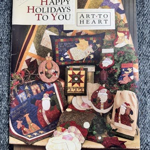 Op de afbeelding: Een boekomslag met de tekst "HAPPY HOLIDAYS TO YOU" en "ART-TO HEART". De omslag toont verschillende kerstgeoriënteerde gequilte items, waaronder de kerstman, engelen en kousen, in rood, groen en beige.