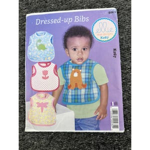 May include: A sewing pattern envelope titled Dressed-up Bibs by Ellie Mae Designs, K267. The purple cover features illustrations of three bibs with dinosaur, tulip, and dragonfly motifs, alongside a photograph of a child wearing a plaid bear bib.