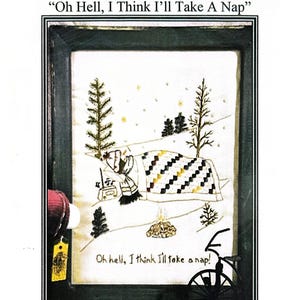 Peut inclure: Image d'une broderie encadrée avec le texte "Oh Hell, I Think I'll Take A Nap". L'œuvre représente une personne sous une couverture dans un paysage enneigé avec des arbres et un petit feu. Le cadre est vert foncé.