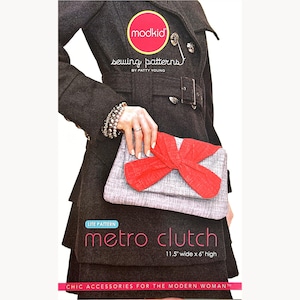 May include: A person holds a grey and red clutch bag with a bow detail. The bag is 29 cm wide and 15 cm high. The text on the bag reads "metro clutch" and "CHIC ACCESSORIES FOR THE MODERN WOMAN". The person is wearing a dark grey coat.