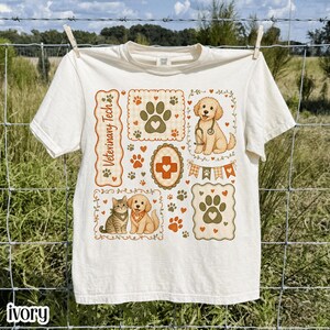 Puede incluir: Una camiseta de manga corta color marfil con un estampado gráfico que incluye el texto Veterinary Tech. El diseño presenta ilustraciones de un perro con estetoscopio, un gato, huellas, corazones y una cruz médica en tonos naranja y verde.